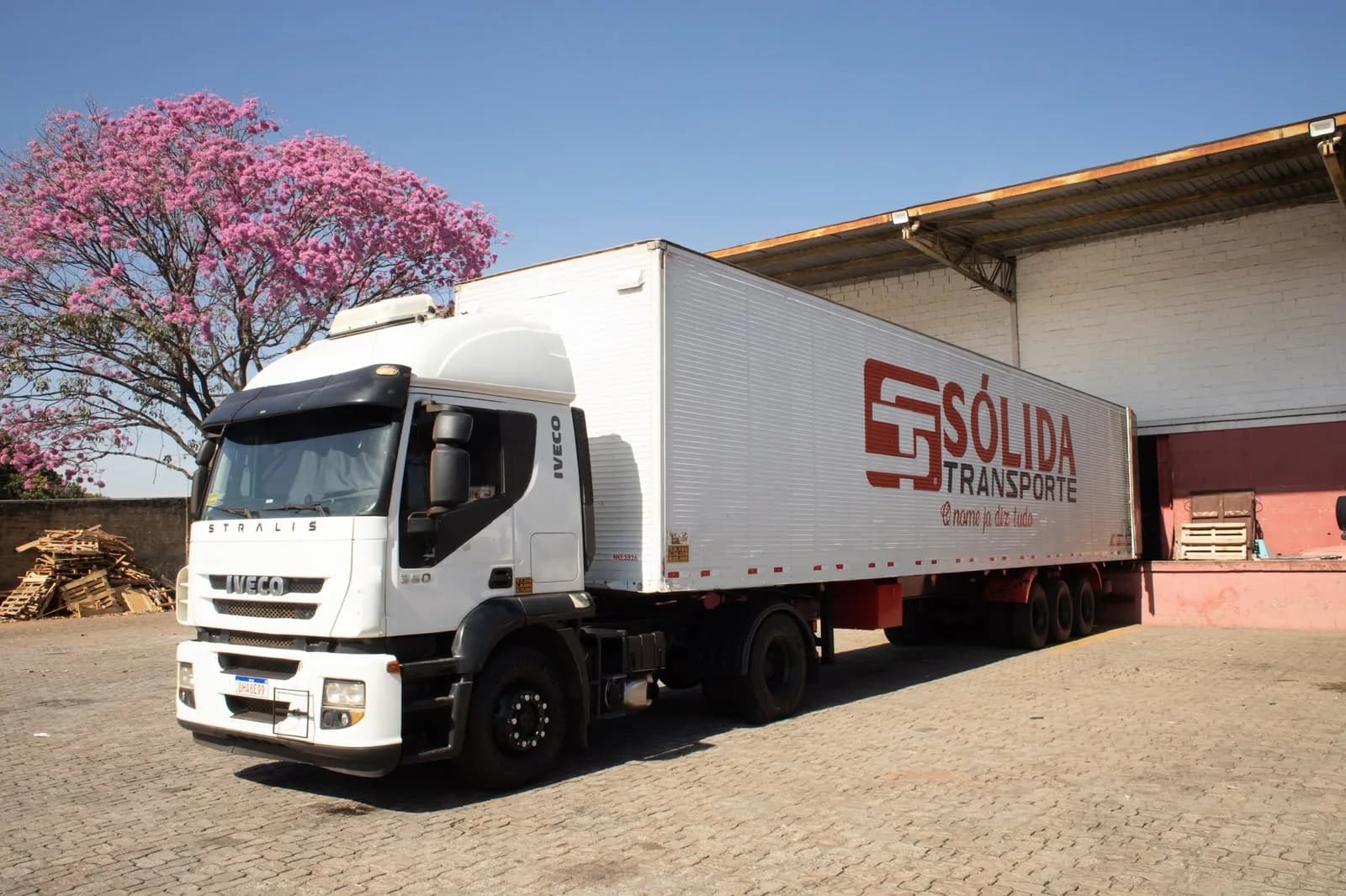 Carreta Iveco Stralis da Sólida encostada na doca do galpão, com um ipê florido ao fundo